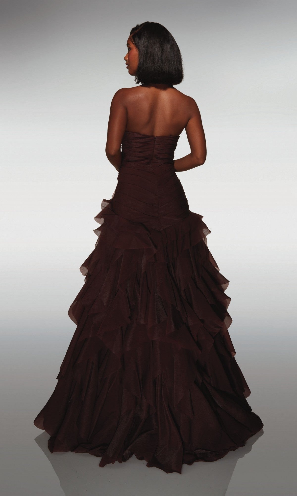 A woman in a strapless, tiered dark brown Alyce 62024 long prom dress stands facing away against a neutral background.